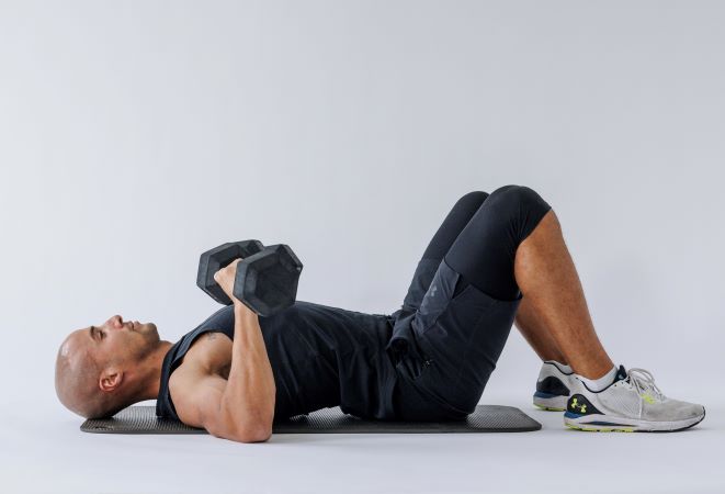 man demonstrates floor chest press: he lays on his back, knees bent, holding two dumbbells at his chest