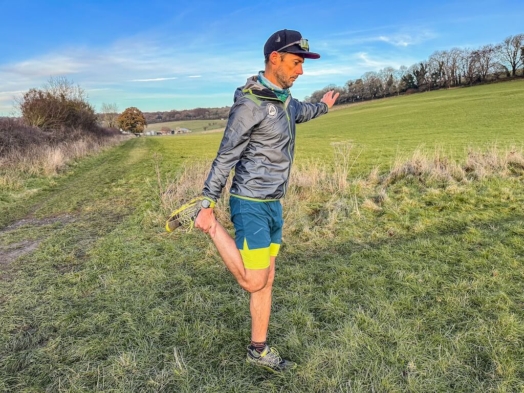 Running coach Simon James performing quads stretch in a field after a trail run