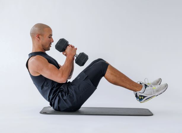 man demonstrates russian twist: balancing on his bottom on the floor, his legs and torso lifted, he holds a dumbbell in two hands at his chest
