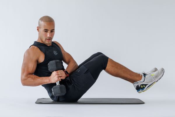 man demonstrates russian twist: balancing on his bottom on the floor, he twists to move a dumbbell either side of him